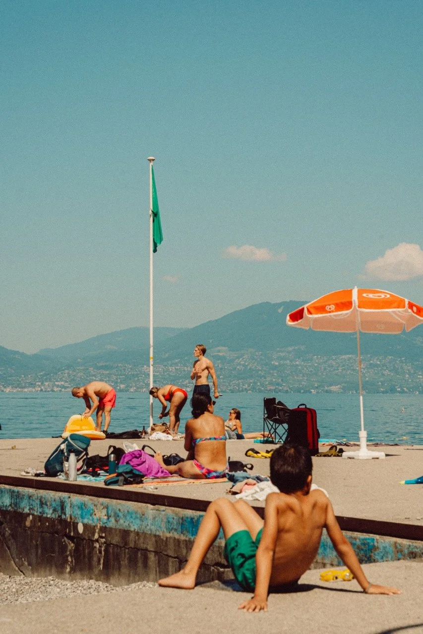People relaxing and swimming at a beach with mountains and a lake in the background. Some are sitting on the shore, others are preparing to swim, and a boy is sitting on the beach near a colorful umbrella and beach gear.