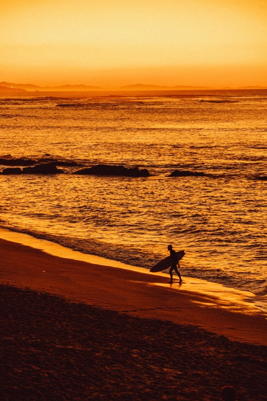 Silhouette of a person carrying a surfboard on a beach during sunset with orange sky and calm water