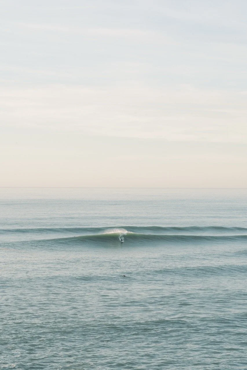 A calm ocean with gentle waves and a surfer riding a wave in the distance under a pale, cloudy sky.