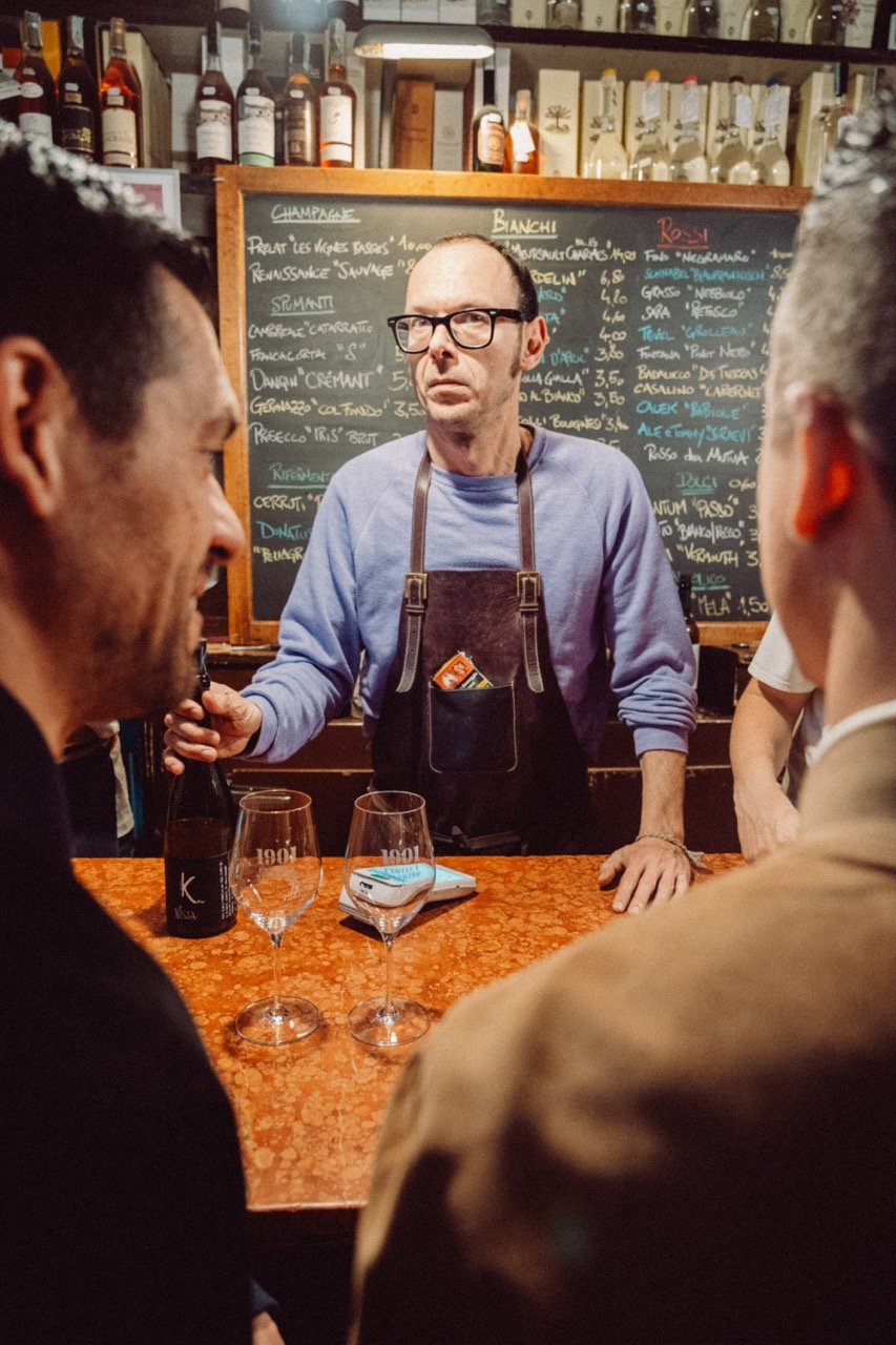 A bartender wearing glasses and a blue shirt standing behind a bar counter, surrounded by customers, with a chalkboard menu in the background listing drinks and prices.