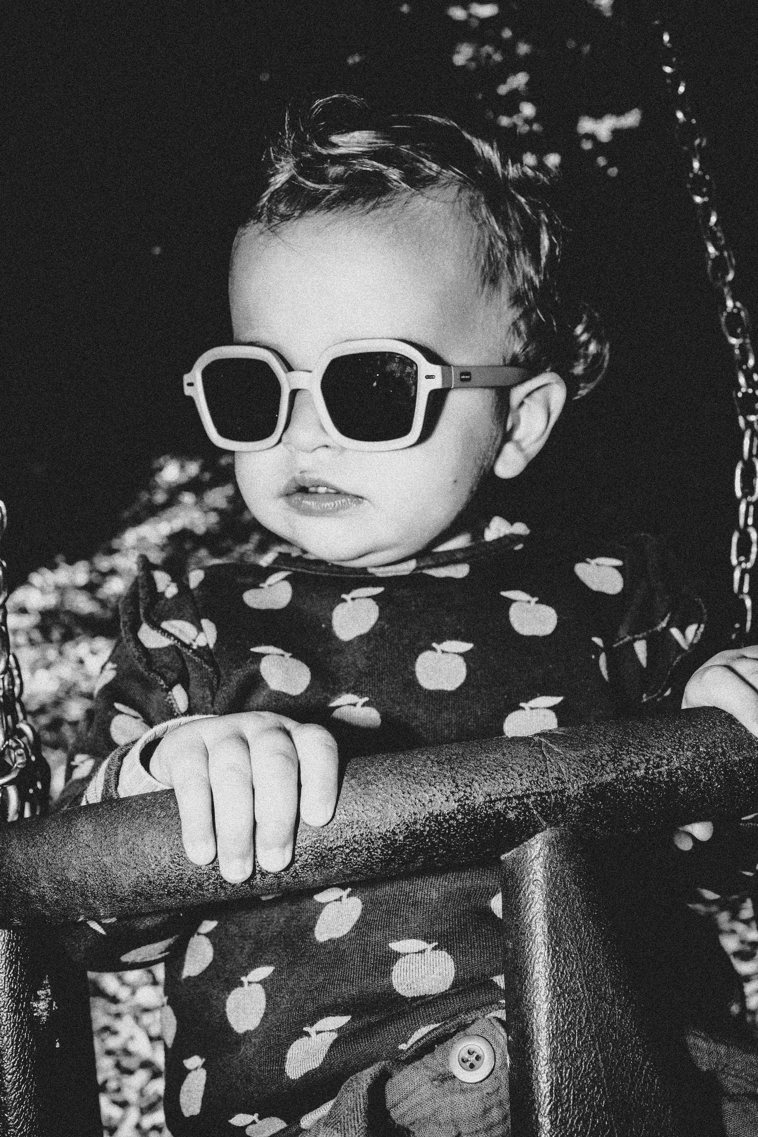 A young child wearing large sunglasses with round lenses and a polka dot outfit, sitting on a swing with a chain and textured seat, outdoors.