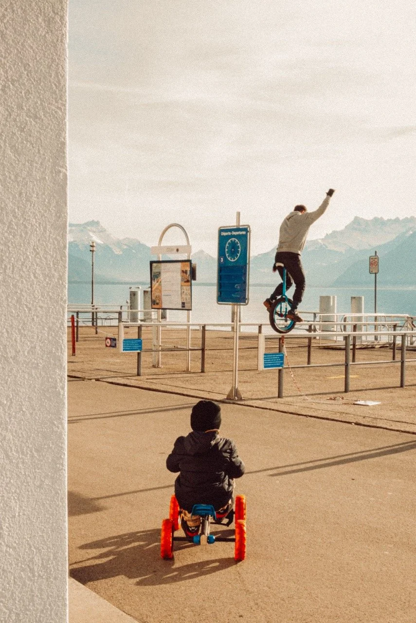 A young boy riding a tricycle on a sidewalk observing a man performing a wheelie on a unicycle, near a waterfront with mountains in the background.