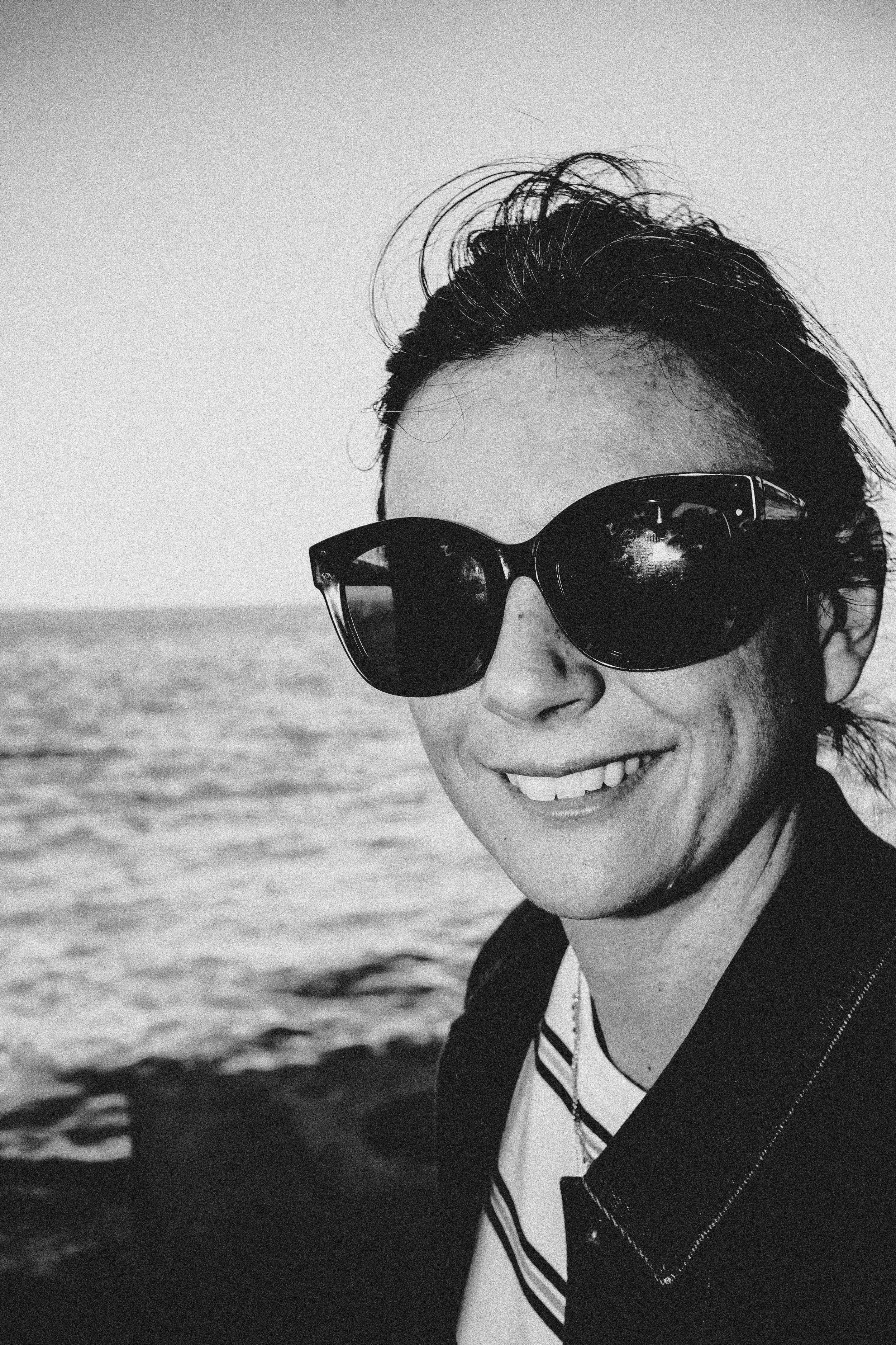 A woman smiling while wearing sunglasses, a striped shirt, and a blazer, outdoors near the water.