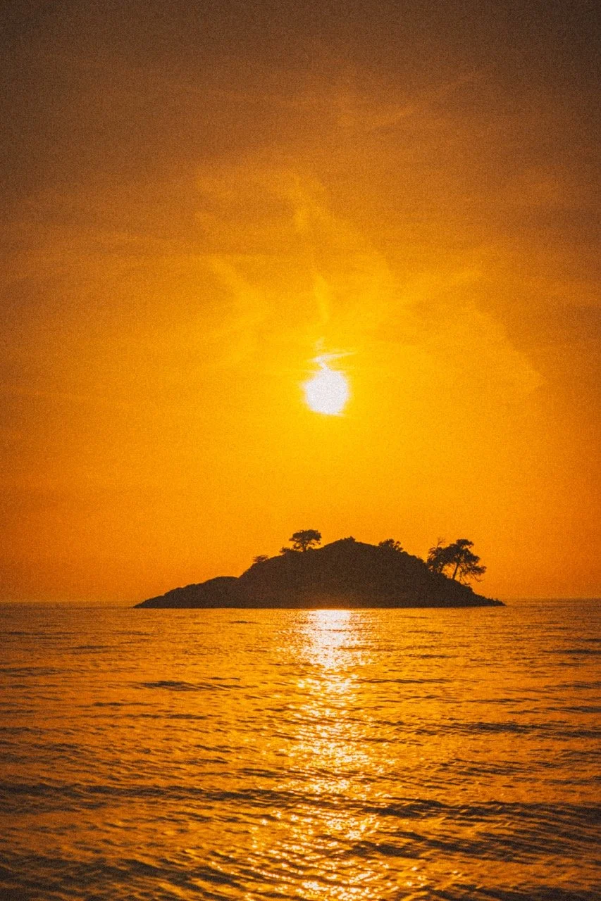 Sunset over an island with silhouettes of trees reflected on the water.