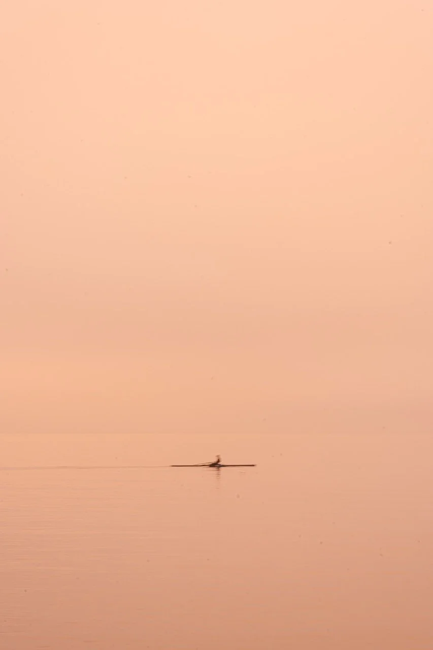 A small boat with a person rowing on calm water during sunset, with a pinkish-orange sky.