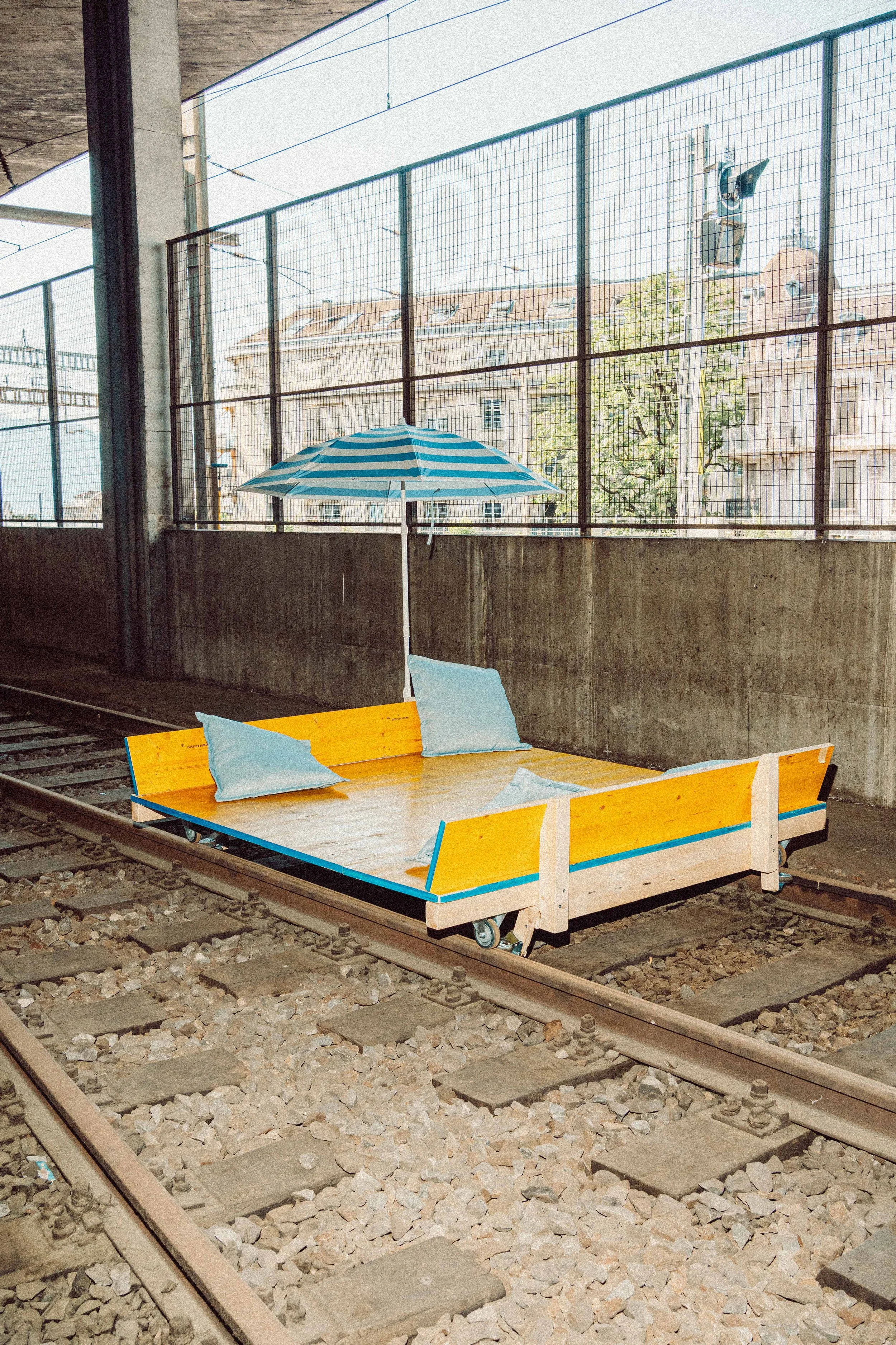 A yellow and blue wooden bench with blue pillows and a blue striped umbrella is placed on train tracks under an overpass, with buildings and a utility pole visible outside.