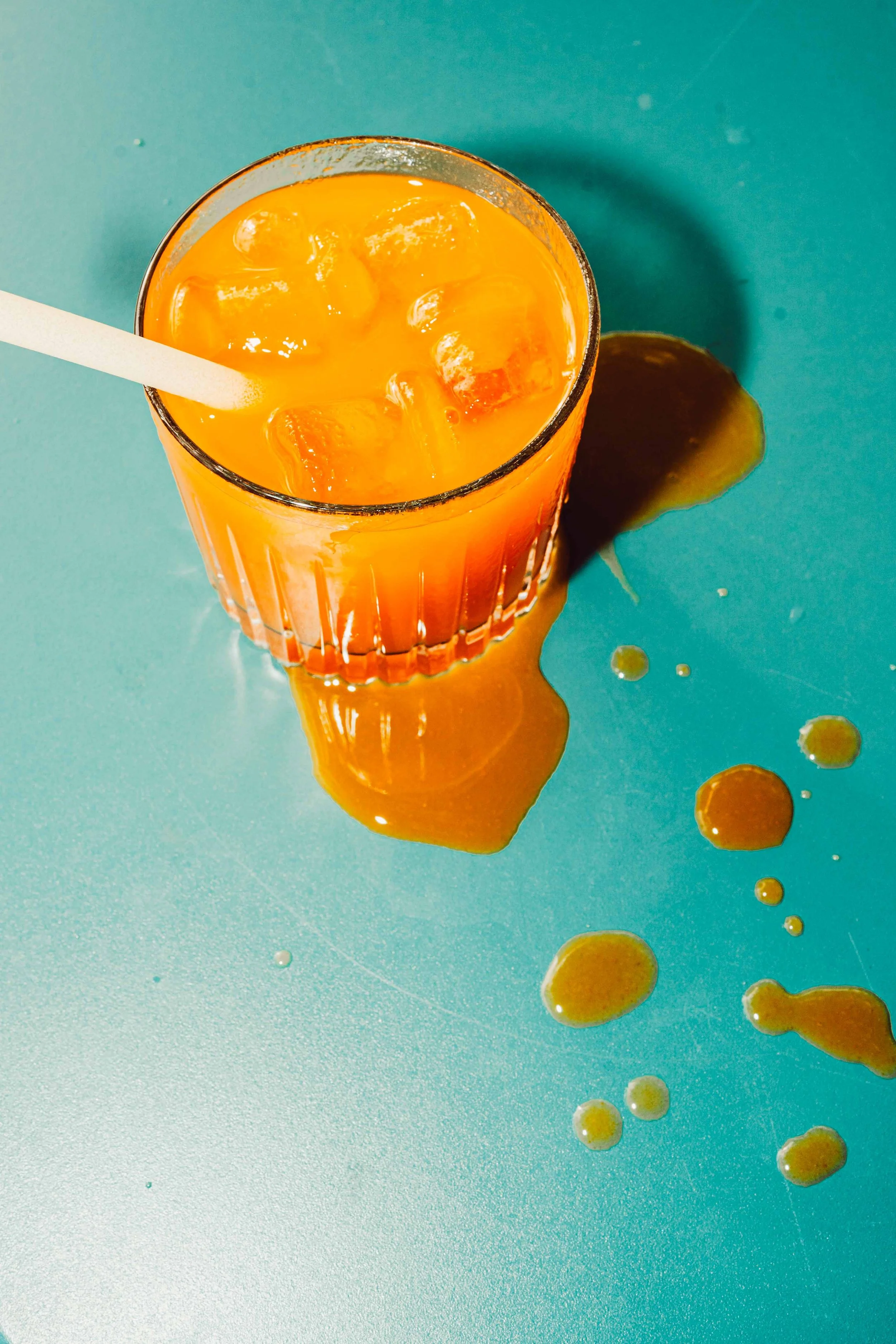 A glass of orange juice with ice cubes on a teal surface, with spilled juice and droplets around the glass.