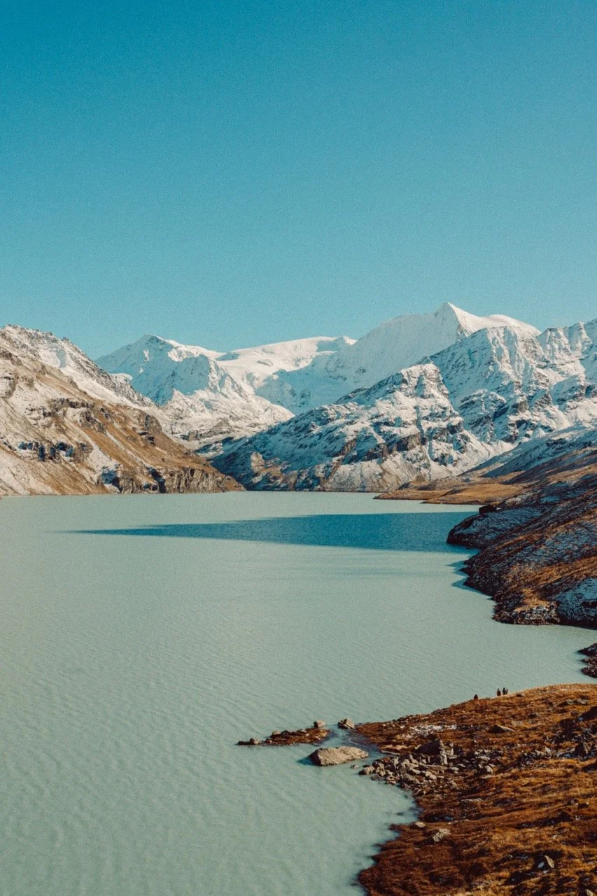 Snow-covered mountains surround a calm, light green lake with rocky shores on the right, under a clear blue sky.