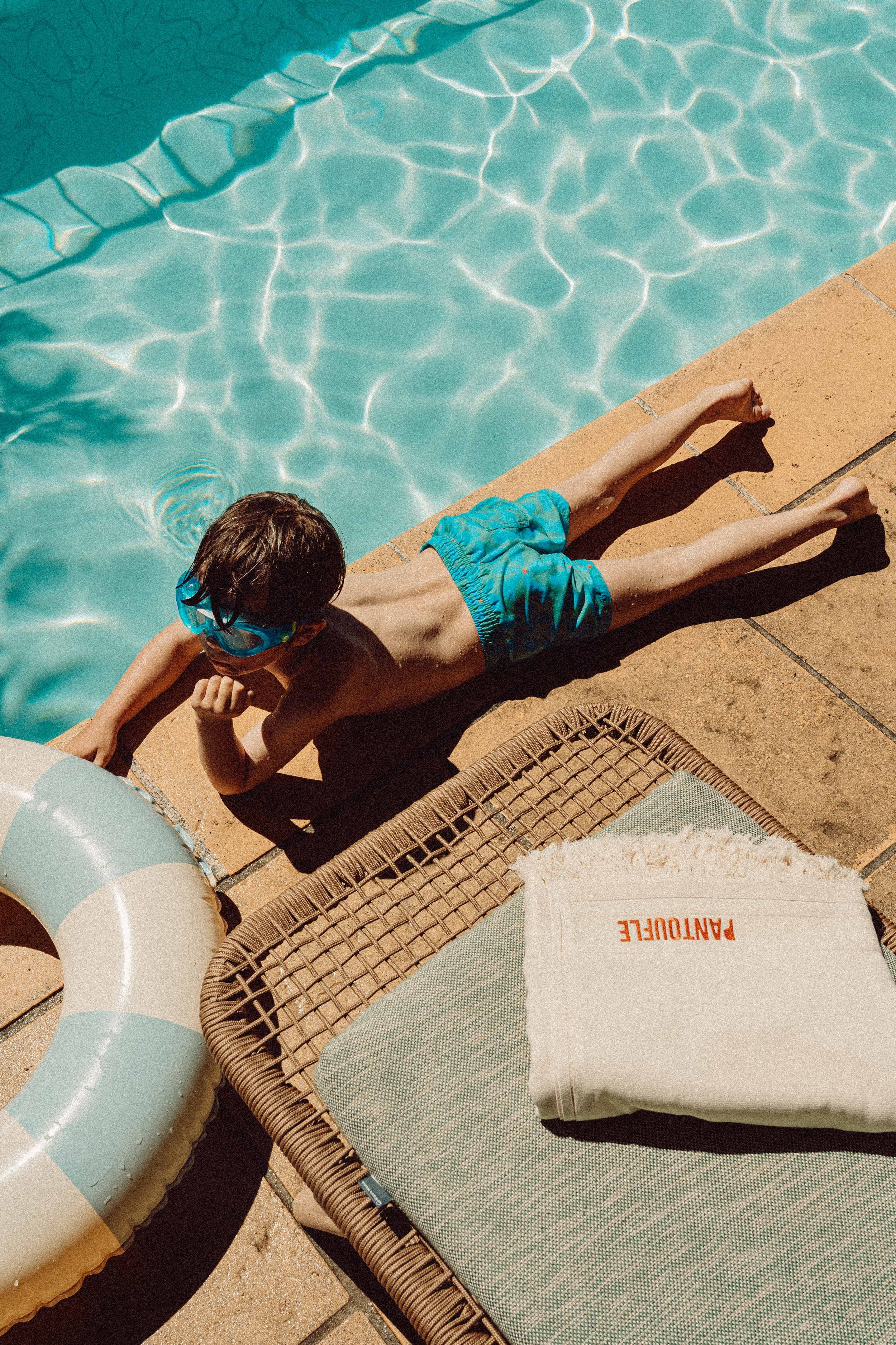 A young boy wearing blue swim trunks and goggles lies on a poolside lounge chair, sunbathing near the edge of a swimming pool.