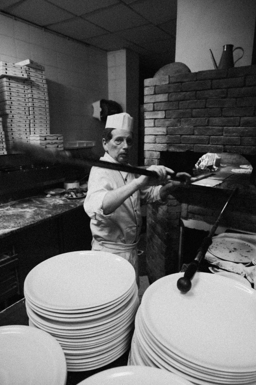 A chef wearing a paper hat is baking pizzas in a brick oven, with stacks of plates on the counter in front.
