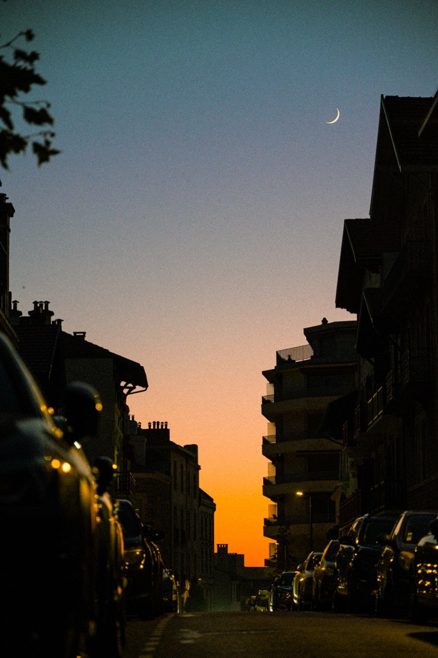 City street at sunset with dark silhouettes of buildings and parked cars, and a slivered crescent moon in the evening sky.