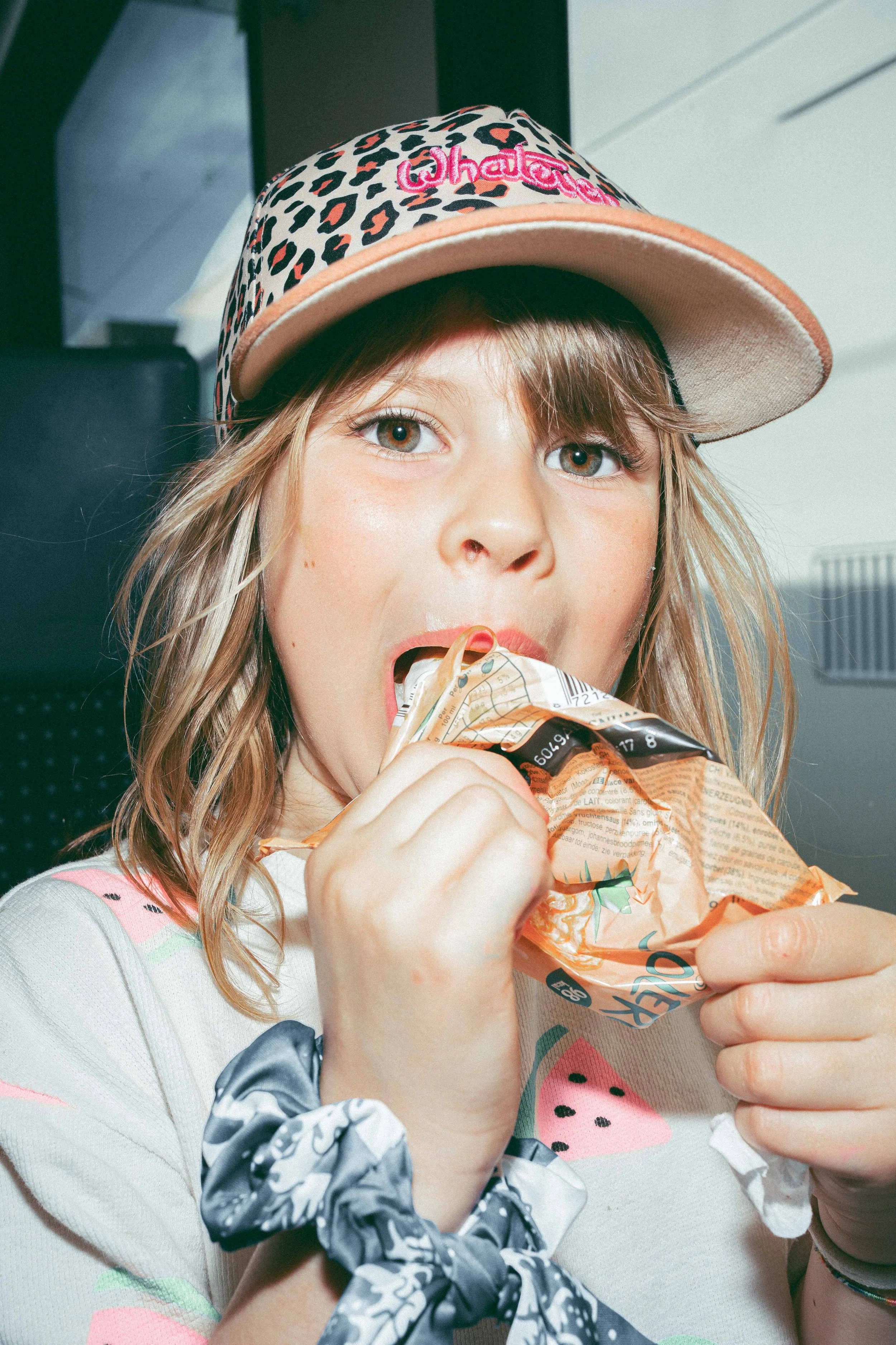 A young woman wearing a leopard print baseball cap with pink embroidery, eating from a snack bag, with a scrunchie on her wrist, and wearing a colorful shirt with watermelon slices and watermelon embroidery.