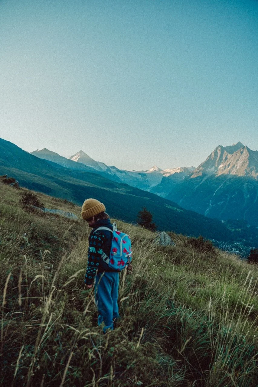 A child wearing a yellow knit beanie, a patterned sweater, and carrying a blue backpack with red and pink designs, hiking through a grassy mountainous landscape.