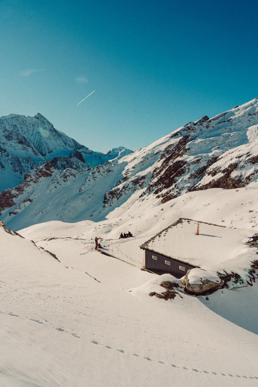 Snow-covered mountain landscape with a small house, people, and ski equipment in the foreground during clear weather.