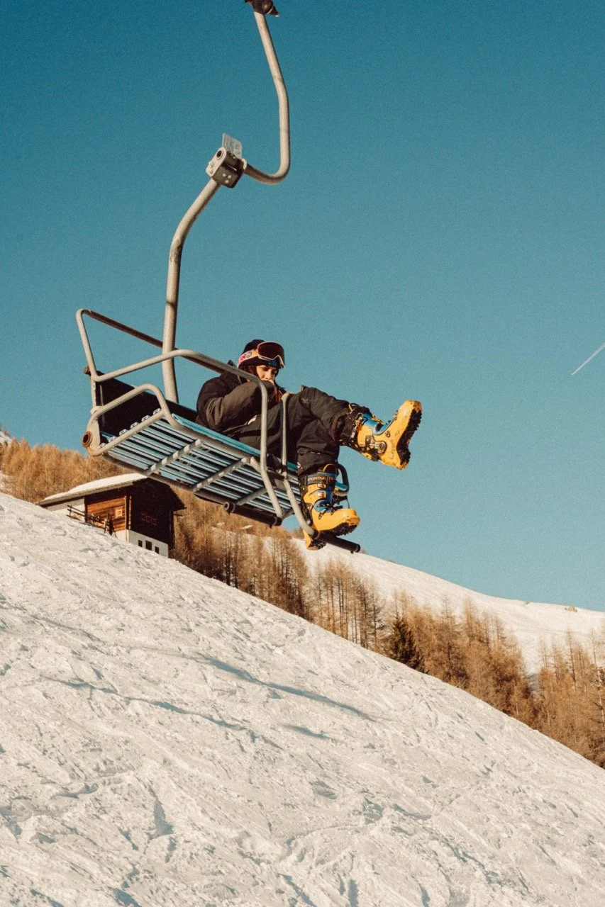 A person sitting on a ski lift chair above snow-covered terrain during daytime, wearing winter gear and yellow ski boots.
