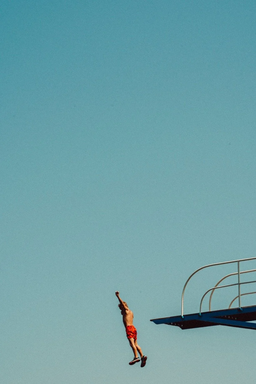 A person in red swim trunks jumping off a diving board into a pool with a clear blue sky in the background.