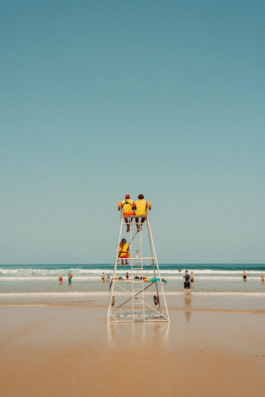 Lifeguards sitting on a lifeguard tower overlooking the beach and ocean, with beachgoers in the water and on the shore.