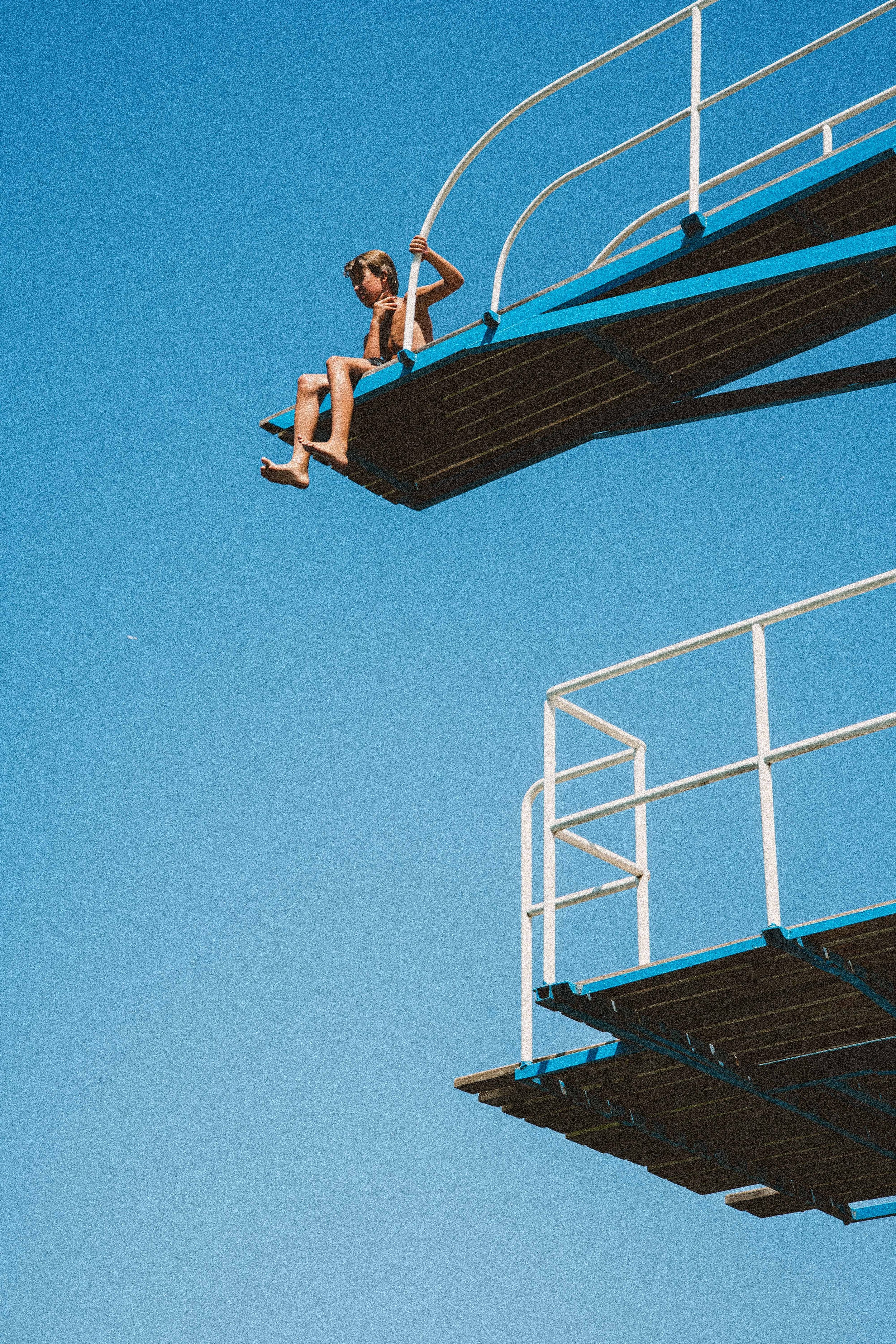 A young boy sitting on the edge of a diving board, looking at a small device in his hand, with a clear blue sky in the background.