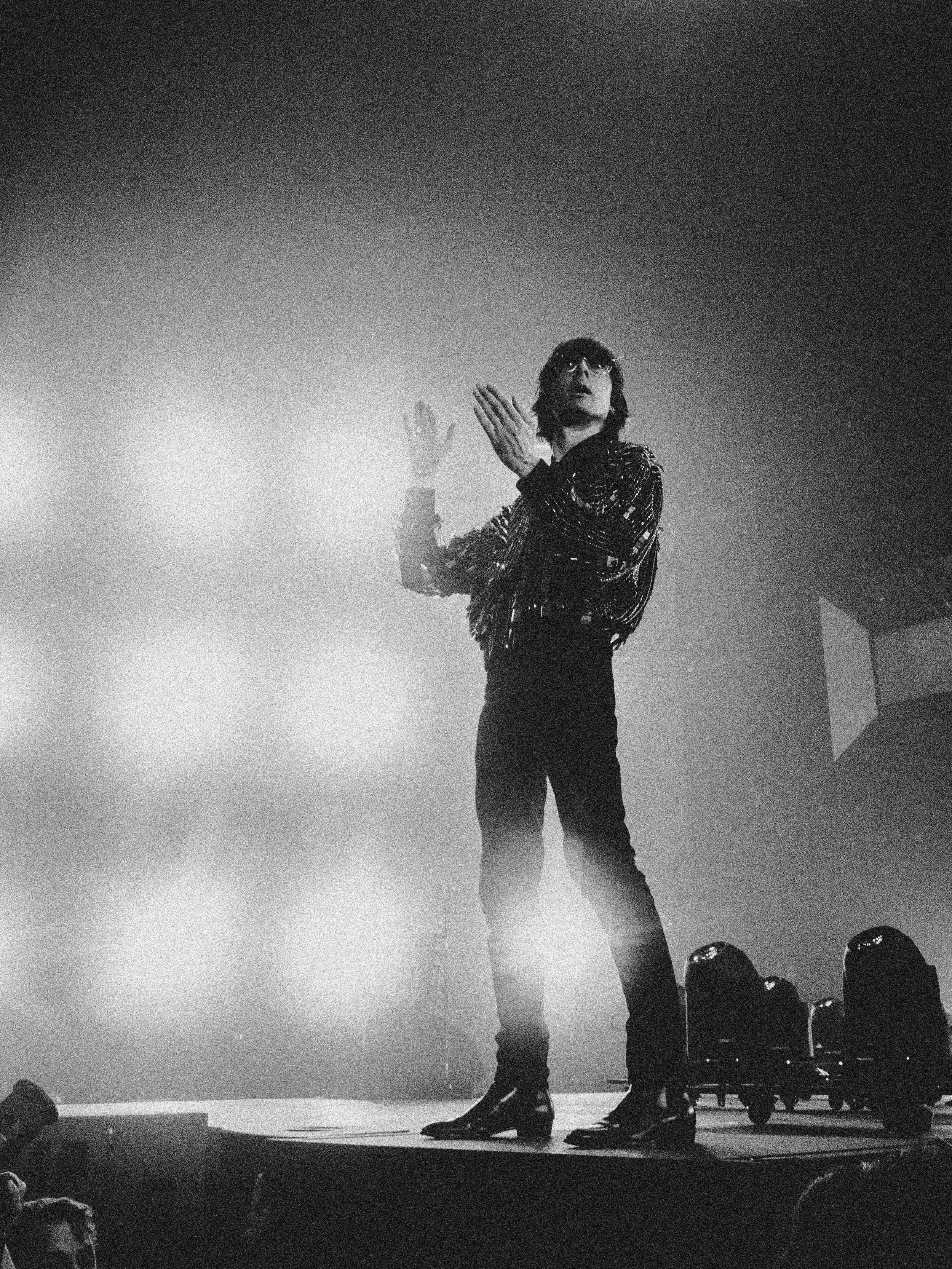 Black and white photo of a performer on stage, standing with hands raised, looking upwards, surrounded by stage lights.