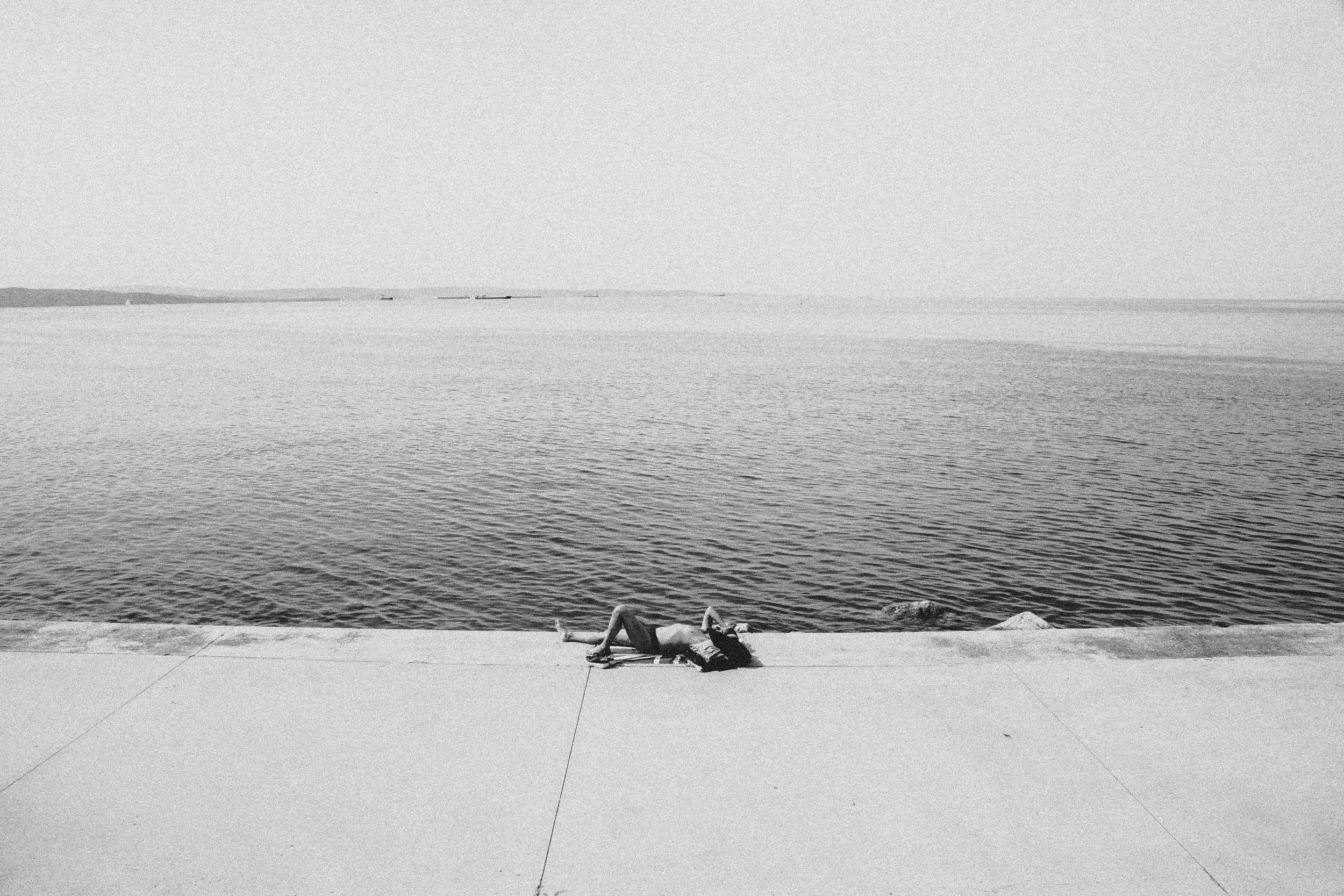 Person lying on the ground near a body of water, with their arm resting on their face, overlooking the water and distant ships on the horizon.
