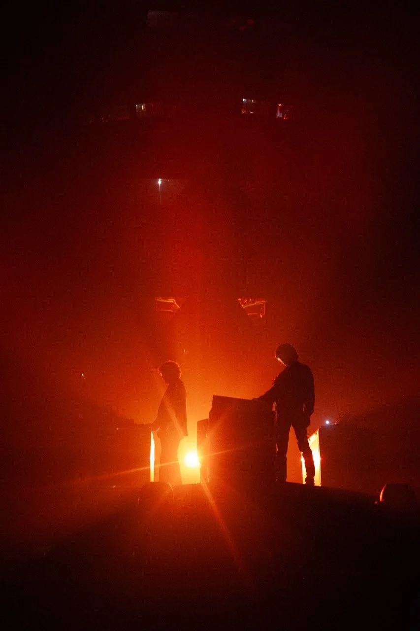 Two people are performing on stage with red lighting and fog, creating silhouettes against the bright background.
