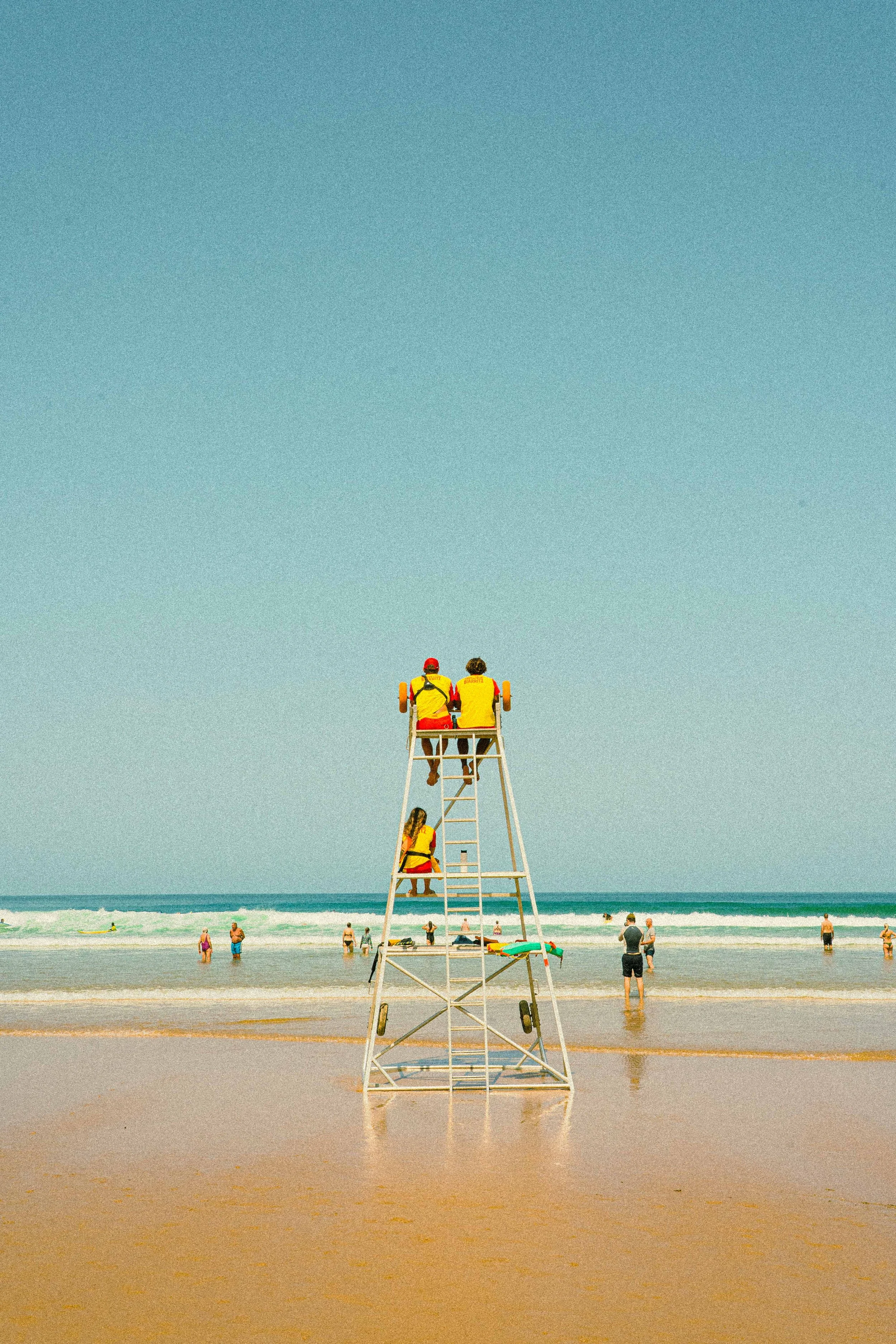View of a crowded beach with a lifeguard tower in the center, two lifeguards sitting at the top wearing yellow shirts, colorful umbrellas, and people swimming, wading, and walking along the shore under a bright blue sky.