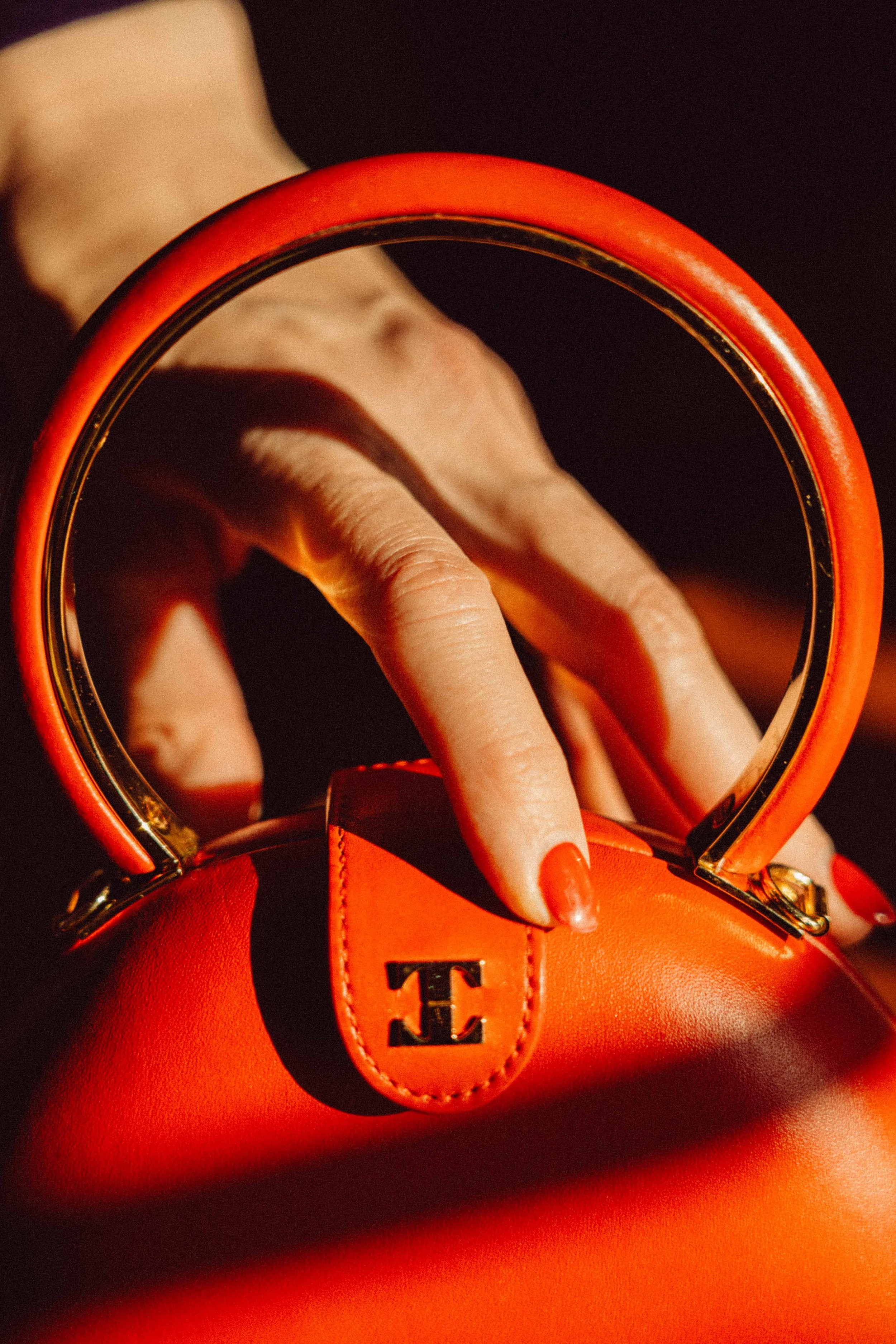 Close-up of a person holding a bright orange handbag with a gold-tone handle and logo tag, with a black background.
