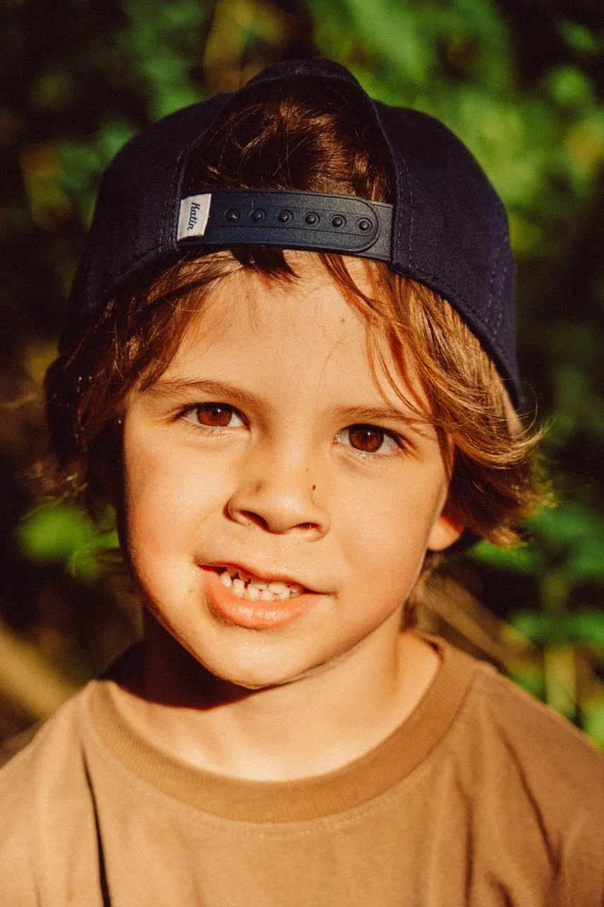 A young boy with brown eyes and brown hair, wearing a black baseball cap backwards and a brown T-shirt, smiling in a natural outdoor setting.