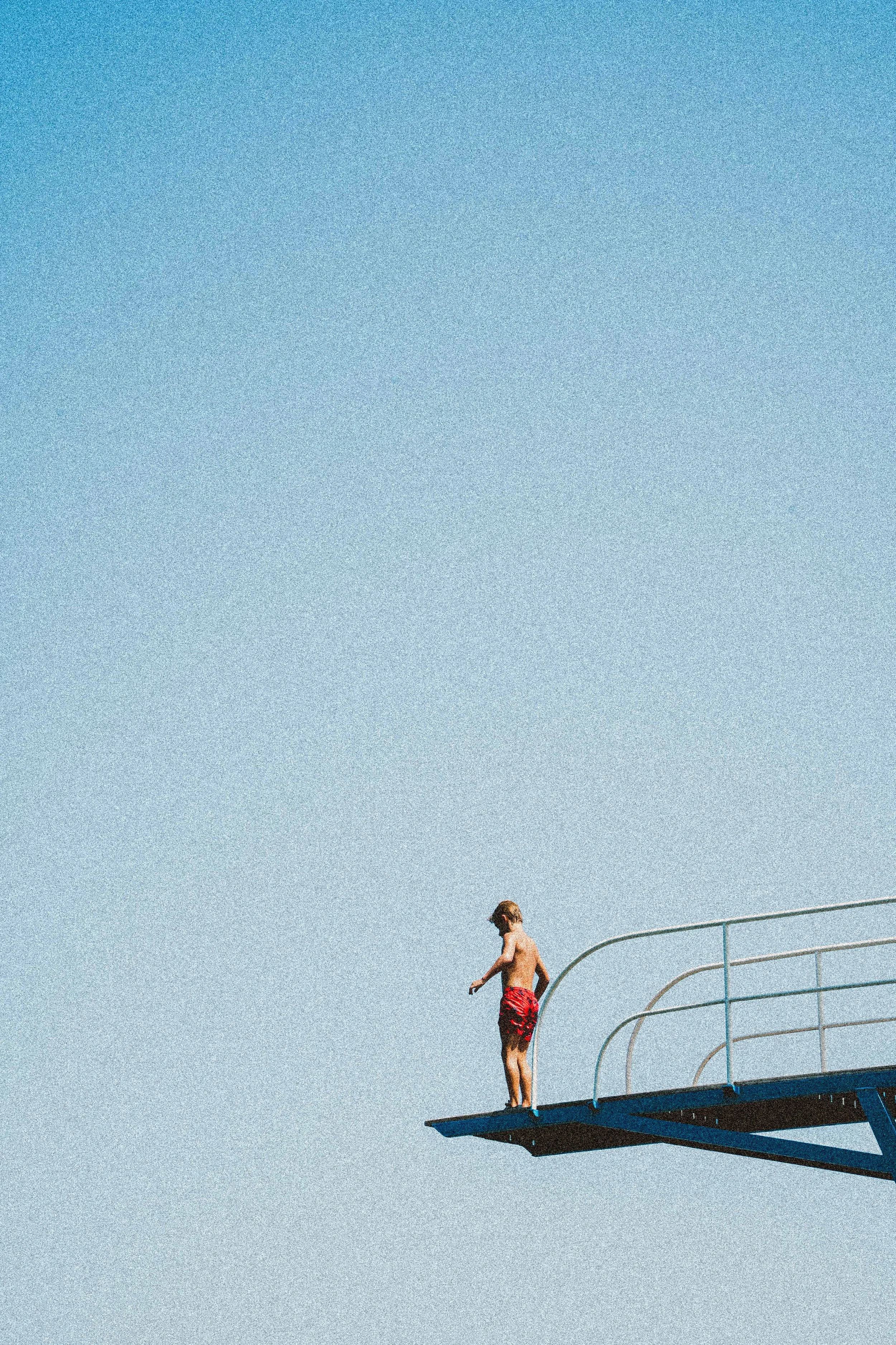 A young boy wearing red swim trunks stands on the edge of a diving board, preparing to jump, with a clear blue sky overhead.
