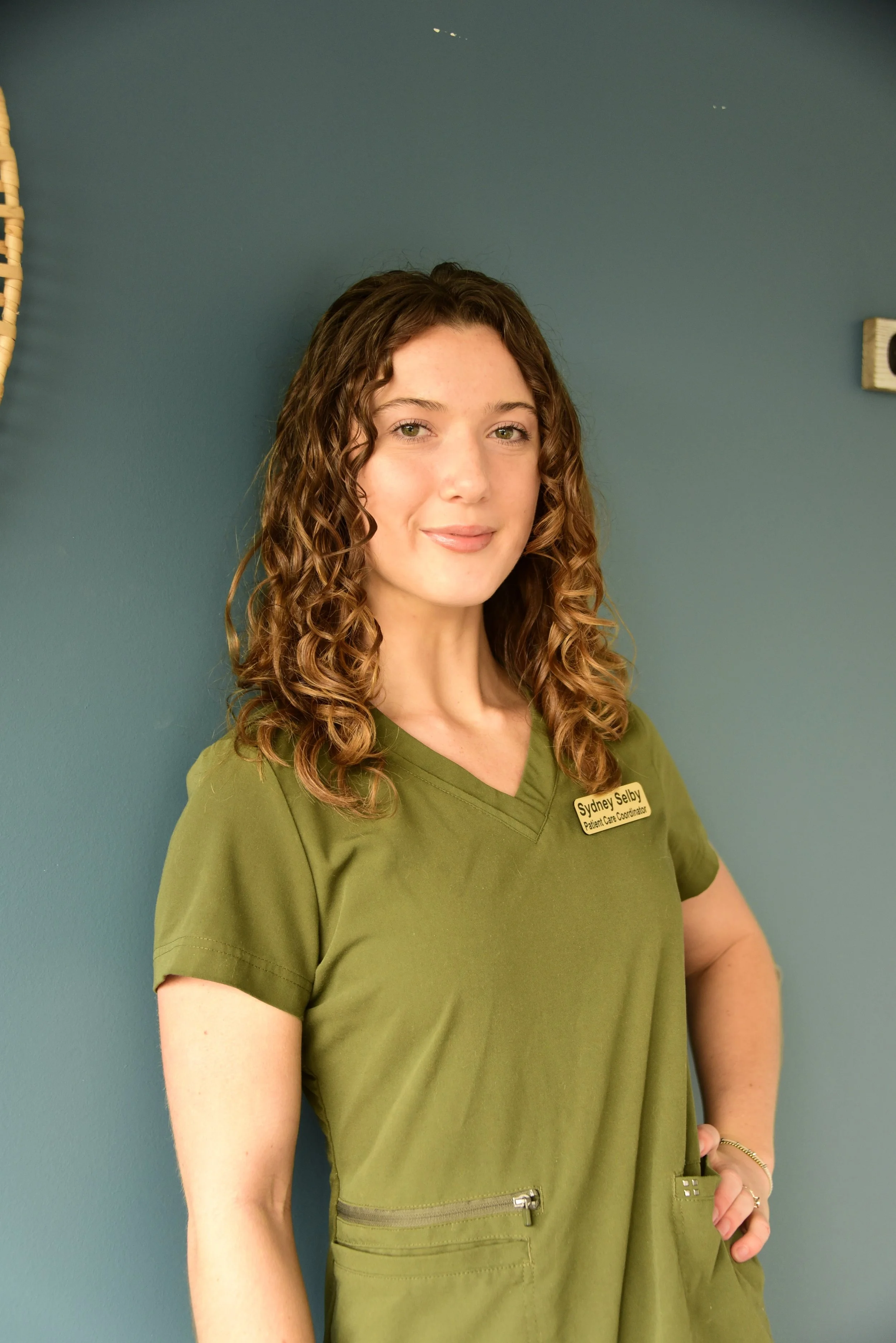 A young woman with curly brown hair wearing green scrubs with a name badge, standing against a teal wall.