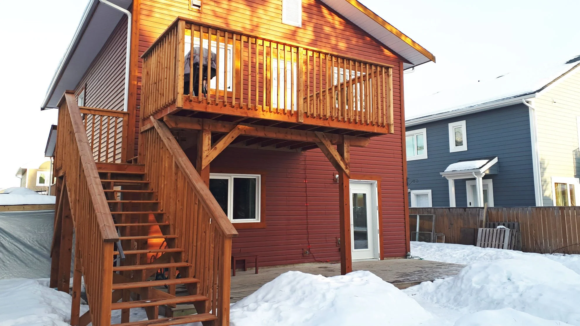 Two-story house with red siding and wooden balcony, surrounded by snow.