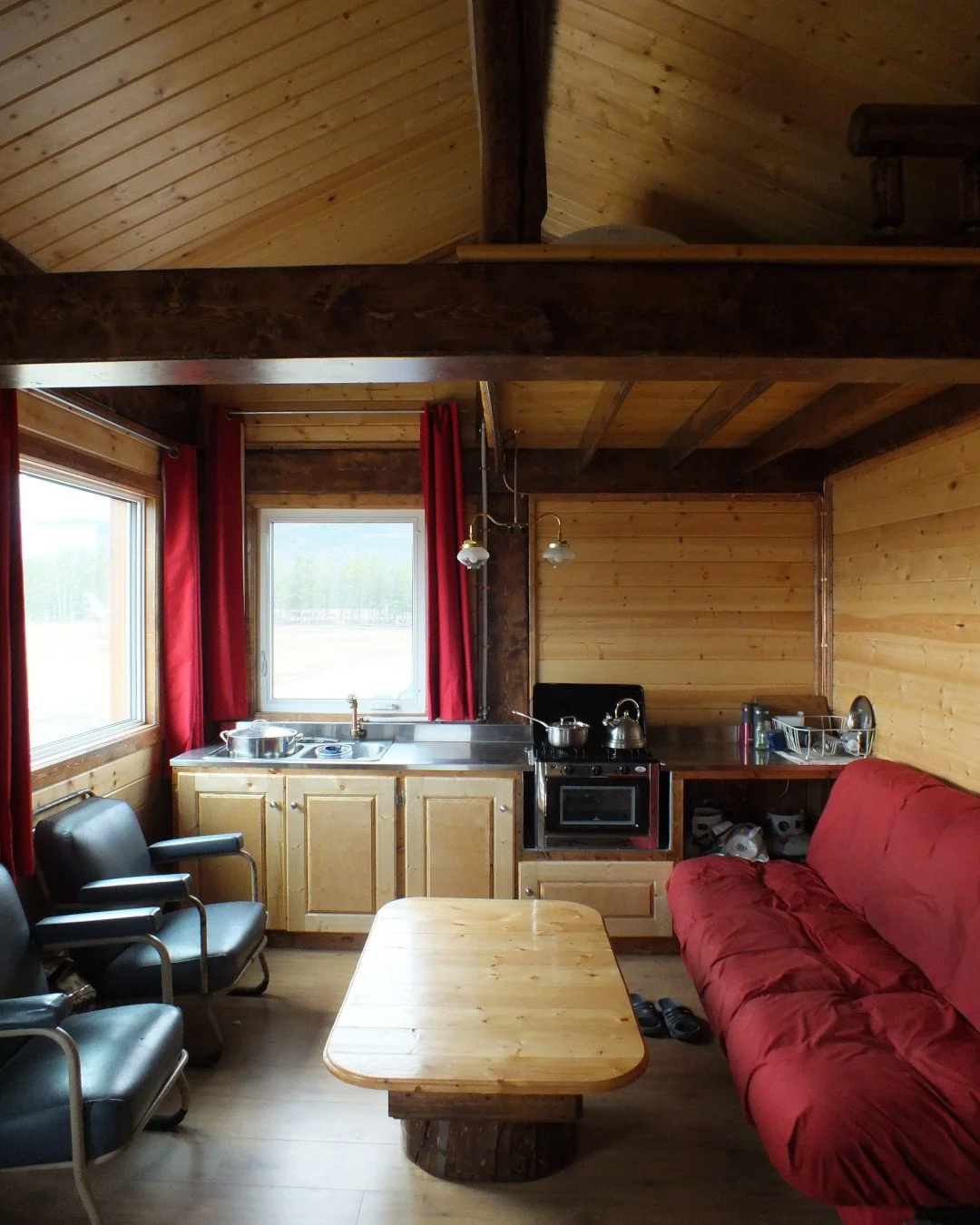 Cozy wooden cabin interior with a kitchenette, red curtains, metal chairs, a red futon, and a wooden coffee table.