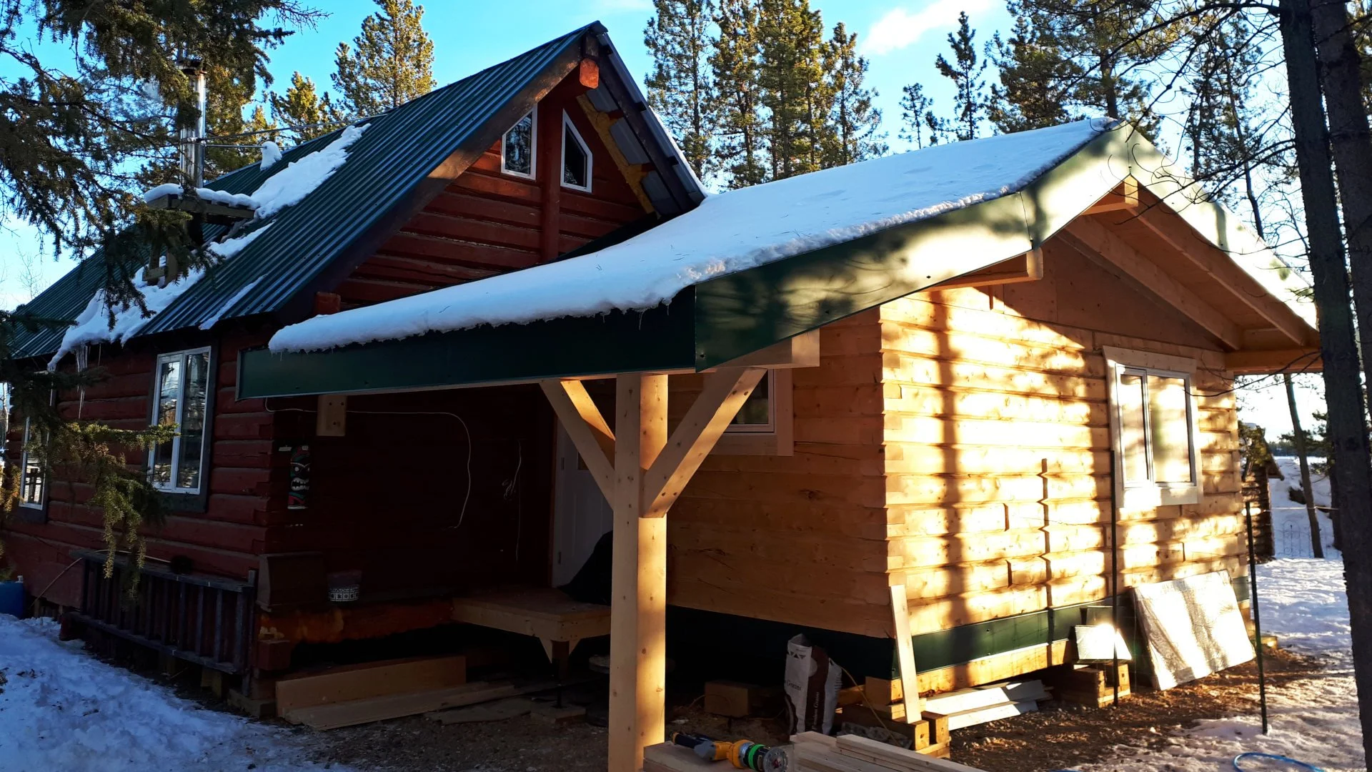 Wooden log cabin with green metal roof and snow, surrounded by trees in a forest setting.