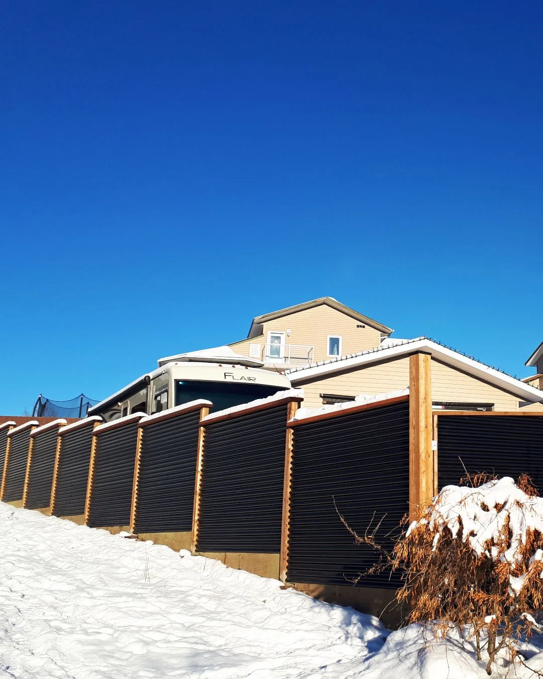 A house with a beige exterior and a black corrugated fence, set against a blue sky. Snow covers the ground and fence, with a small bush visible in the foreground.