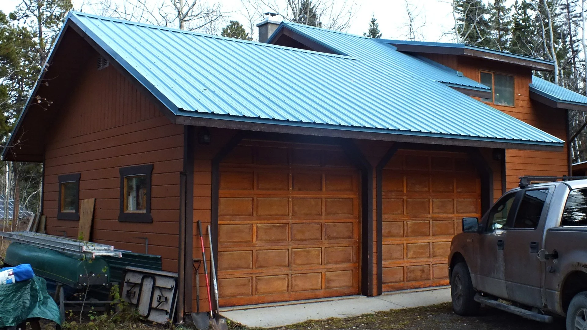 Two-car garage with brown wooden doors and a blue metal roof, adjacent to a pickup truck and assorted tools.