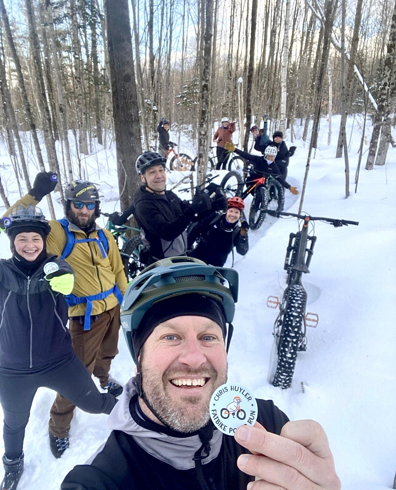 Group of people in winter gear standing with mountain bikes in a snowy forest, smiling and posing for a selfie, with one person in the foreground holding a badge that reads 'Chris Huyler Fatbike Poker Run'.