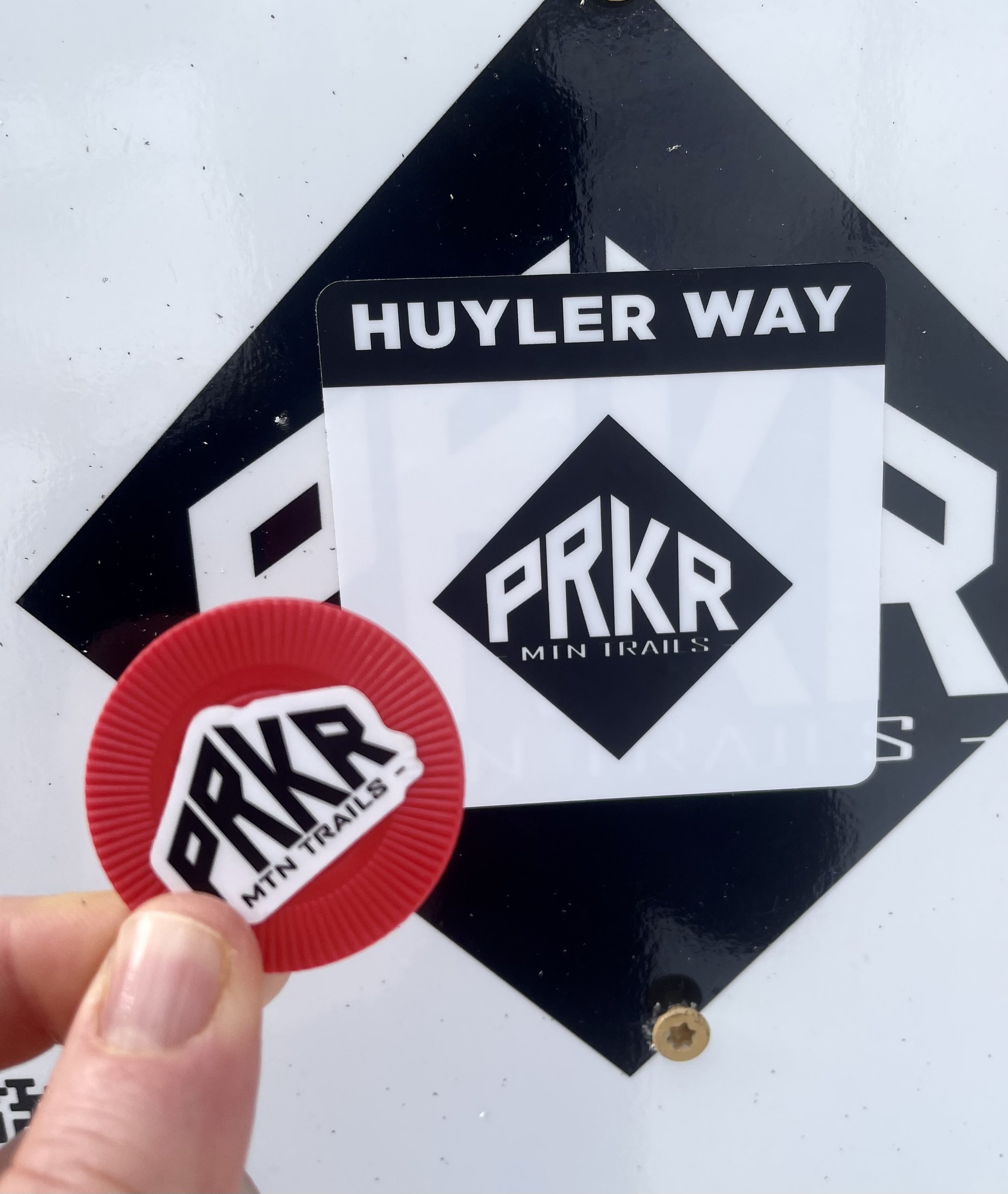 A hand holding a red badge with the logo 'PRKR MTN TRAILS' in front of a white surface featuring a black and white sticker with the 'PRKR MIN TRAILS' logo, a black border, and the text 'HUYLER WAY' at the top.