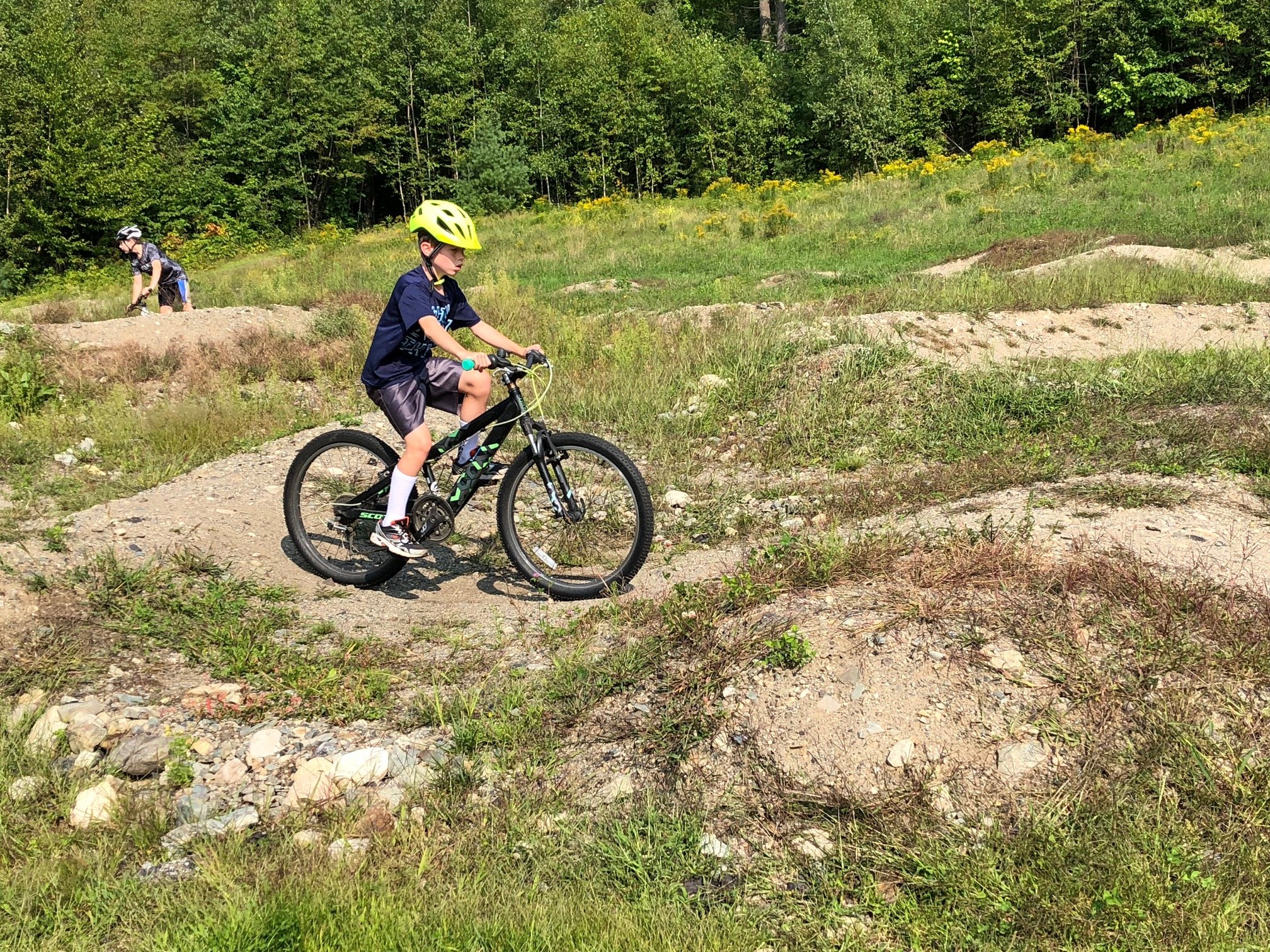 Two boys riding bikes on a dirt trail in a grassy area surrounded by trees.