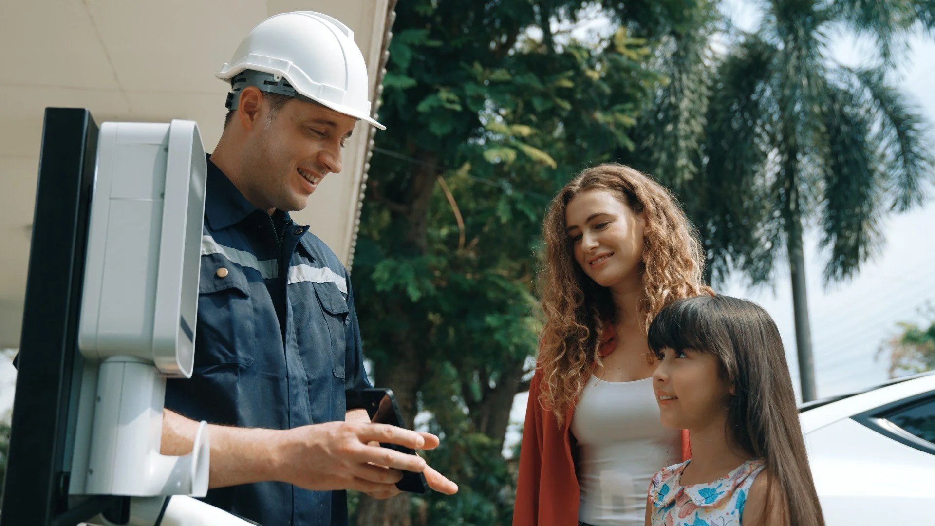 Ein Mitarbeiter in blauer Uniform und weißem Schutzhelm zeigt den Familienmitgliedern auf einem Smartphone eine Information, während die Familie, bestehend aus Mutter und zwei jungen Mädchen, interessiert zuschaut. Sie stehen im Freien mit Bäumen und einem Auto im Hintergrund.