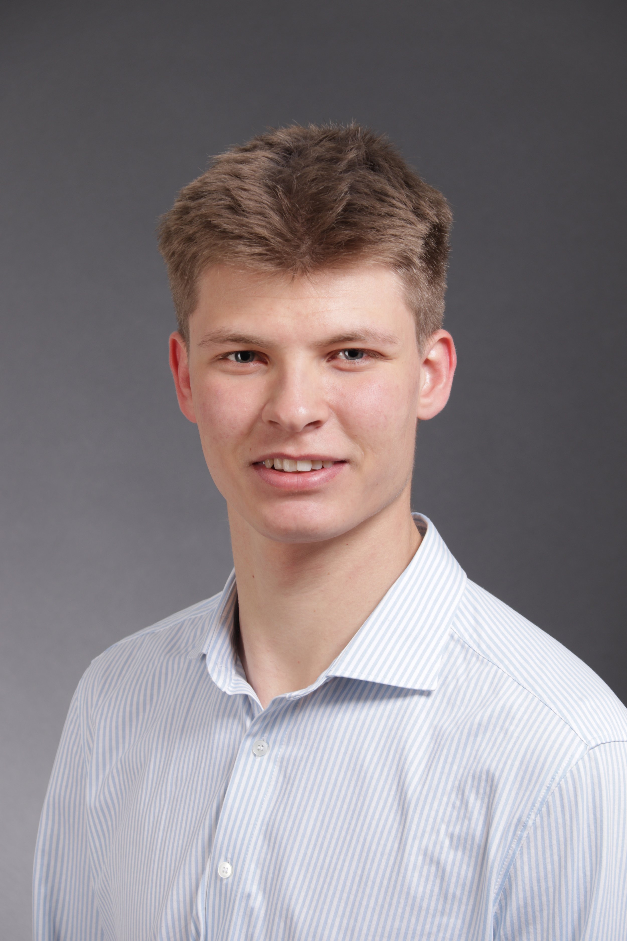 Porträt eines jungen Mannes vor grauem Hintergrund, trägt ein weiß-blau gestreiftes Hemd, lächelt leicht.
