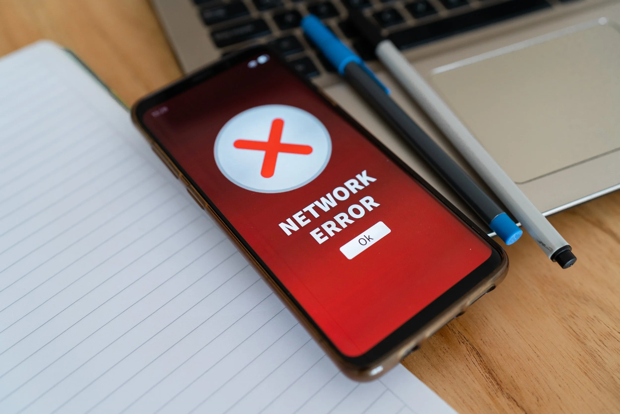 Smartphone auf einem Tisch mit einem Fehlerbildschirm, der einen Netzwerkfehler zeigt, umgeben von Stiften, einem Laptop und einem Notizbuch.