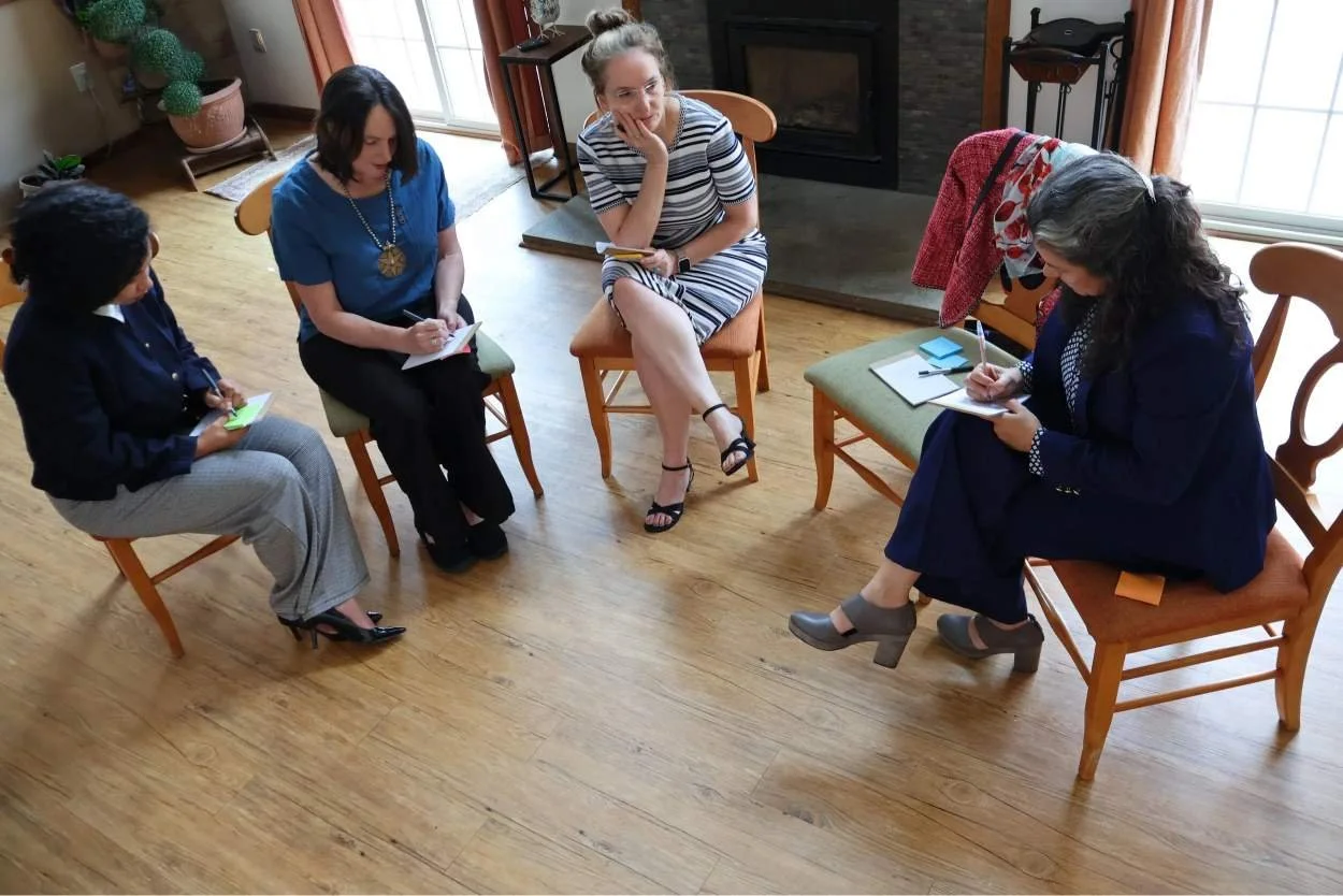 Five women sitting on chairs in a circle, engaged in a conversation, with notebooks and pens, in a cozy room with wooden floors and large windows.