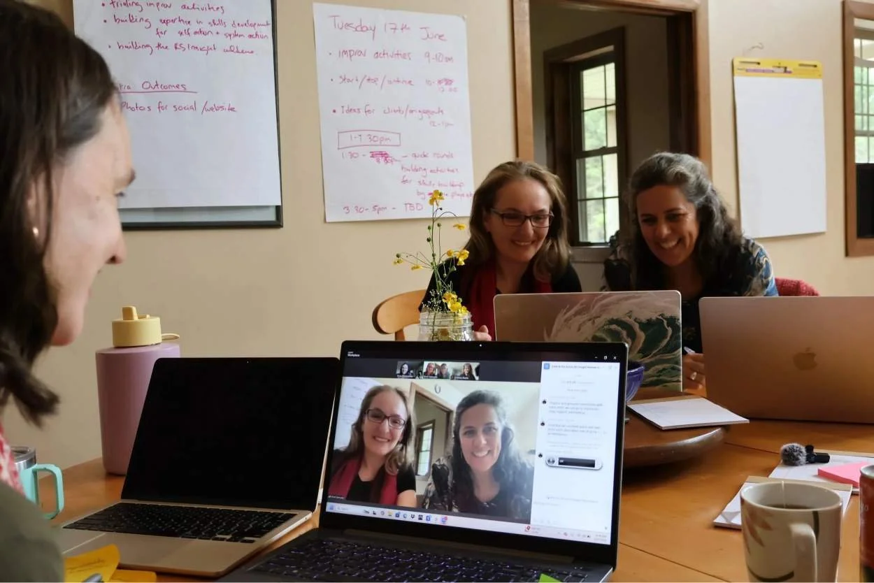 Group of women sitting around a table in a meeting room, some working on laptops, others engaging in a video call, with whiteboards and windows in the background.