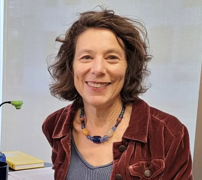 A smiling woman with short, curly brown hair wearing a multicolored beaded necklace, a blue top, and a red jacket, standing in front of a plain light gray background.
