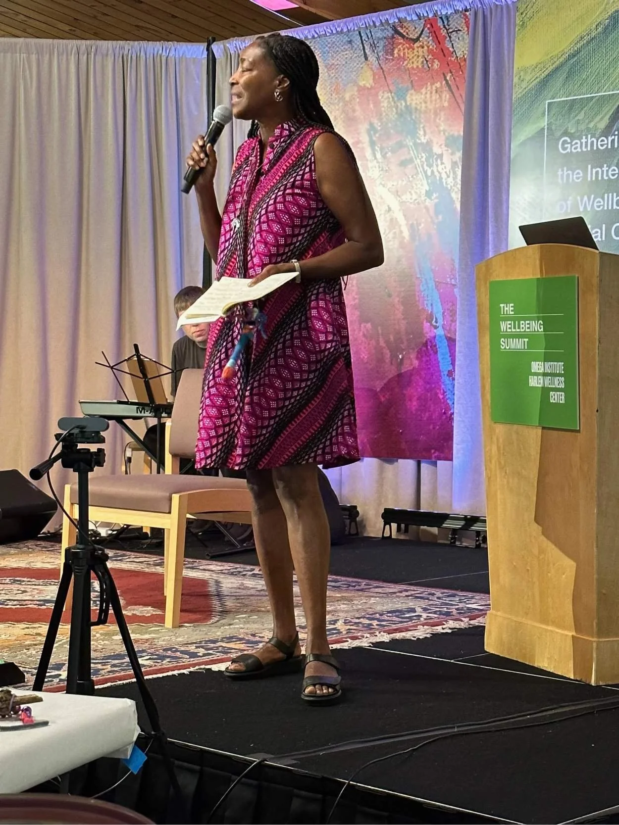 A woman with braided hair wearing a pink and purple patterned dress, standing on a stage, speaking into a microphone while holding a notepad. Behind her, a man is playing a keyboard. The backdrop has colorful abstract art, and there is a wooden podium with a green sign that reads "The Wellbeing Summit" and "Omega Institute Parlen Wellness Center."