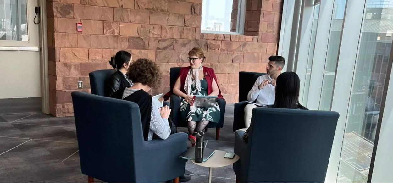 A group of five people sitting in a circle in a modern office lounge area, engaged in conversation, with large windows and cityscape view in the background.