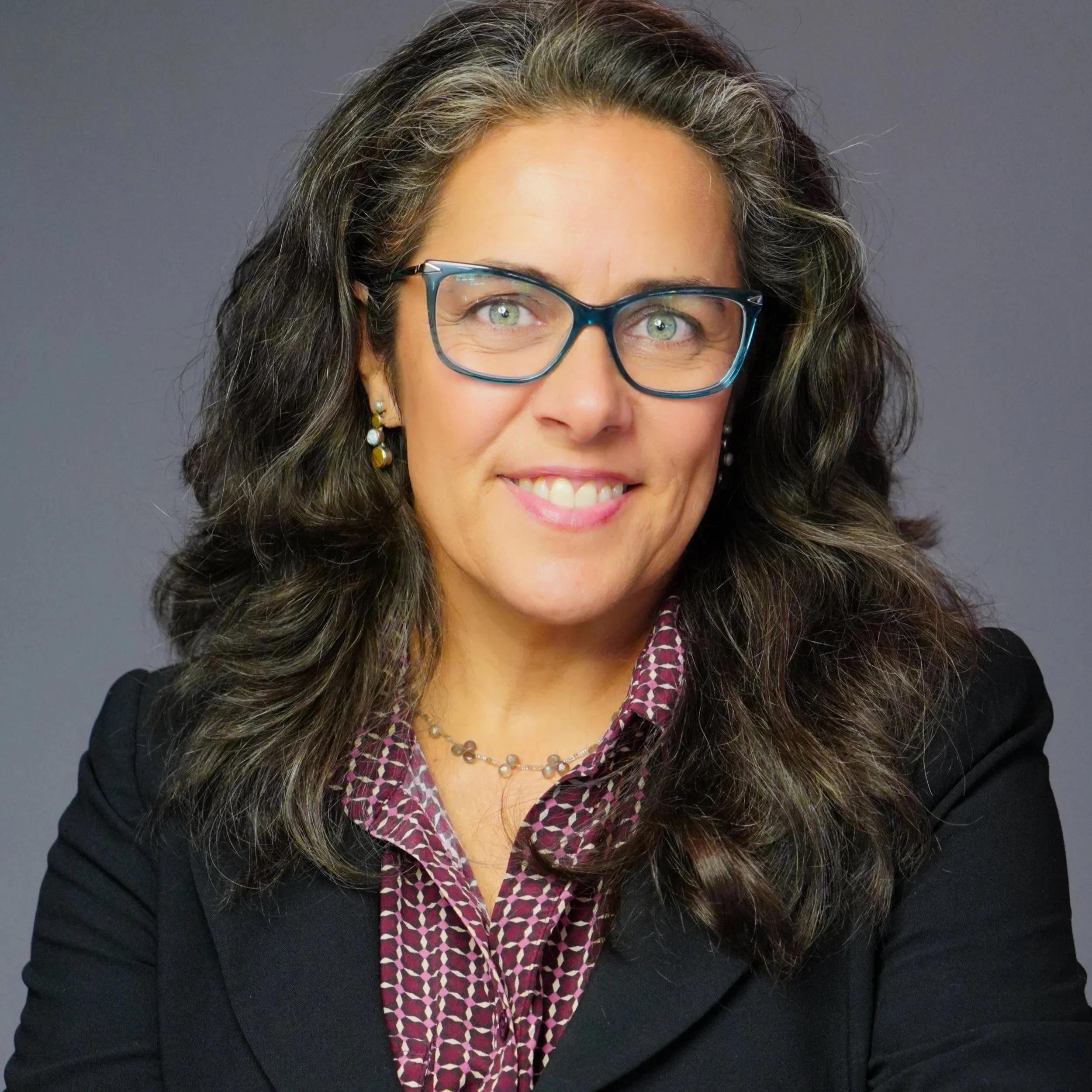 Portrait of a smiling woman with curly brown hair, wearing glasses, a black blazer, a patterned blouse, and jewelry, against a gray background.