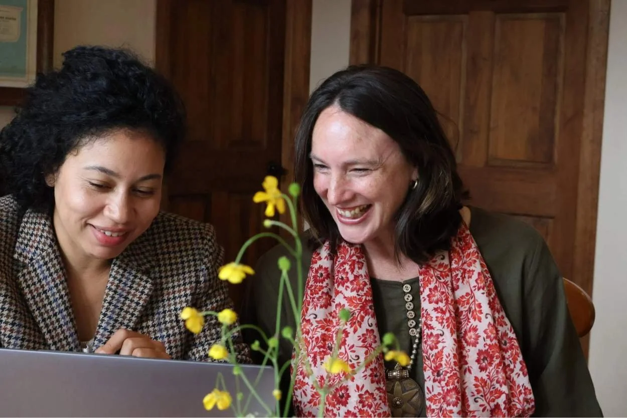 Two women smiling and looking at a laptop, with a small yellow flower in the foreground.