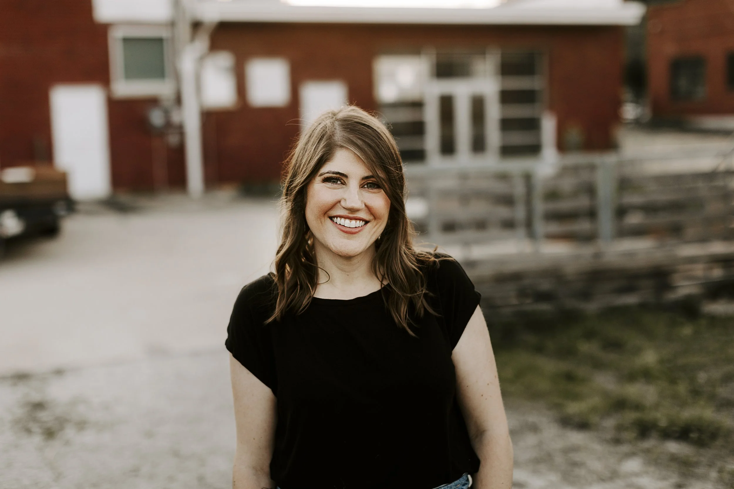 A young woman with long brown hair and a black shirt smiling outdoors near a fence with a red brick building in the background.