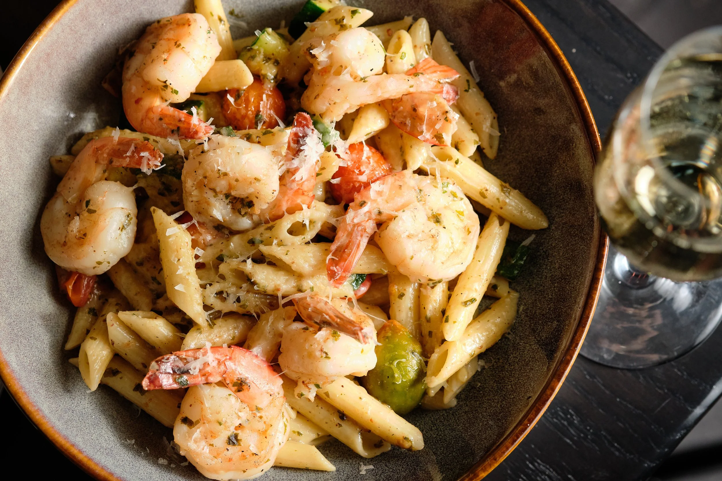 A bowl of shrimp and pasta with herbs, grated cheese, and a side of white wine.